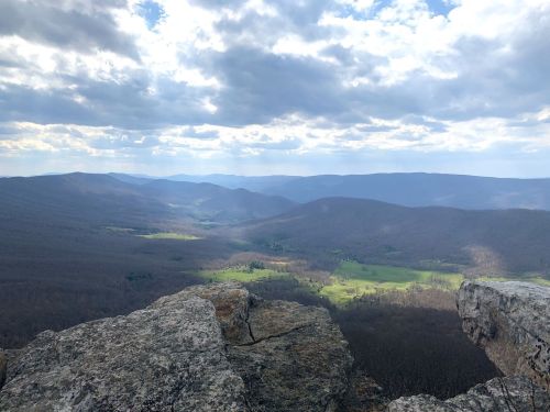 A panoramic view of rolling mountains and a green valley under a cloudy sky, seen from a rocky outcrop.