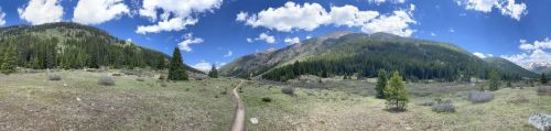 A panoramic view of a lush green valley surrounded by mountains under a bright blue sky with scattered clouds.