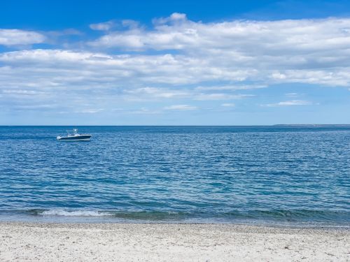A calm beach scene with a boat on the water under a blue sky with scattered clouds.