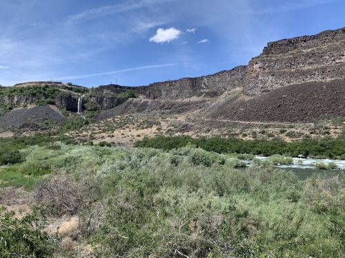A scenic view of a river flowing through lush greenery, with rocky cliffs and a waterfall in the background under a blue sky.