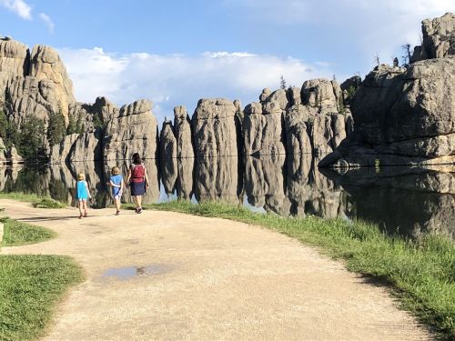 Two children and an adult walk along a path by a calm lake, surrounded by towering rock formations and trees.