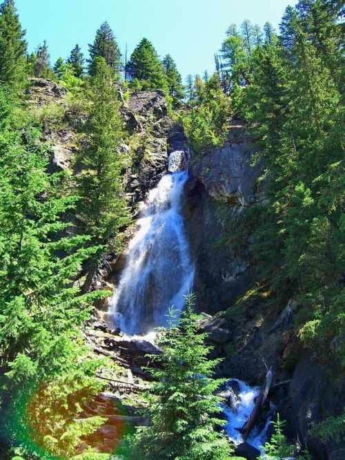 A cascading waterfall flows down rocky cliffs, surrounded by lush green trees under a clear blue sky.