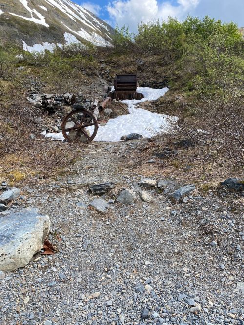 A rocky path leads to a wooden structure with a wheel, surrounded by snow and greenery in a mountainous area.