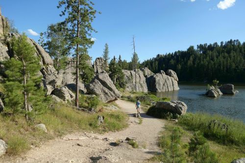 A person walks along a sandy path by a serene lake, surrounded by tall rocks and lush green trees.
