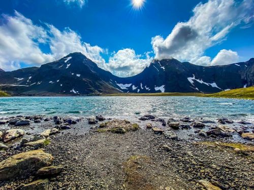 A serene mountain landscape with a clear blue lake, rocky shore, and snow-capped peaks under a bright sky.