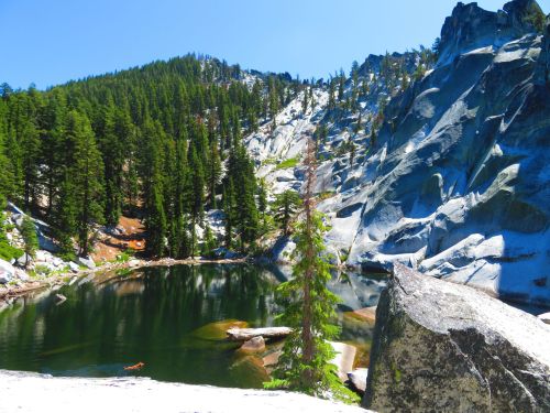A serene mountain landscape featuring a clear lake surrounded by lush green trees and rocky cliffs under a bright blue sky.