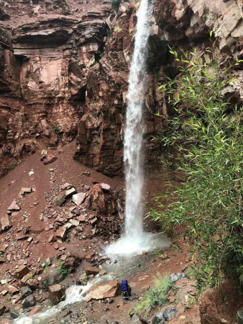 A waterfall cascades down rocky cliffs into a small pool, surrounded by earthy tones and greenery.