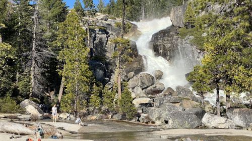 A scenic waterfall cascades over rocks, surrounded by tall trees and people enjoying the natural landscape.