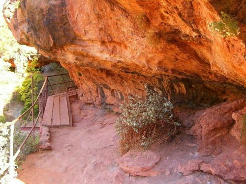 A rocky cliff with reddish-brown stone and a wooden pathway leading into a natural area with sparse vegetation.