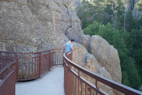 A person walks along a curved wooden railing path beside large rock formations and trees in a natural setting.