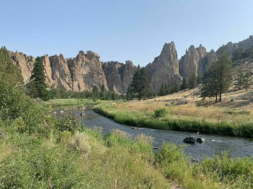 A serene river flows through a grassy landscape, framed by towering rock formations and scattered trees under a clear sky.