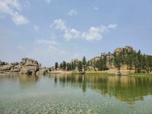 Scenic view of a calm lake surrounded by rocky formations and pine trees under a blue sky with scattered clouds.