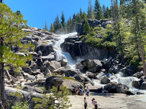 A waterfall cascades over rocky terrain, surrounded by lush trees and people enjoying the scenic view on a sunny day.