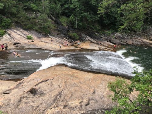 A rocky river area with people swimming and relaxing near a waterfall surrounded by lush greenery.