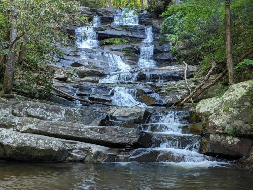 A serene waterfall cascades over rocky steps, surrounded by lush greenery and trees.