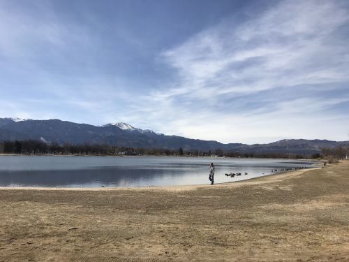 A person walks along a sandy shore by a calm lake, with mountains and a clear sky in the background.