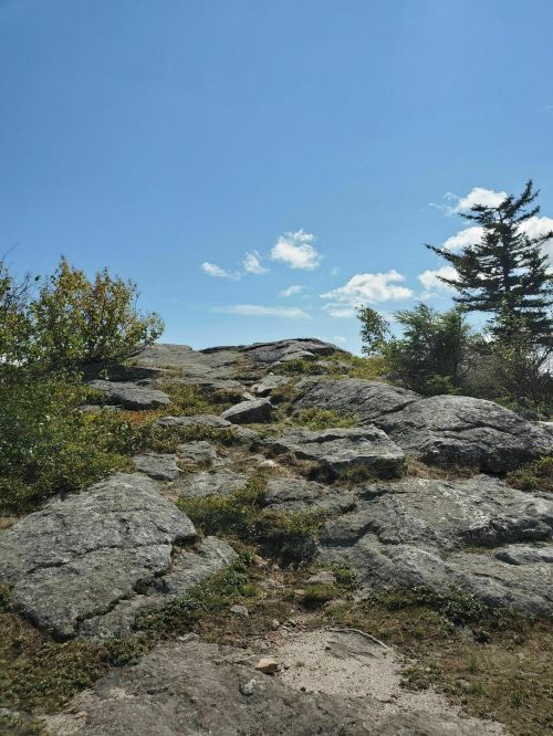 A rocky path leads to a summit under a clear blue sky, surrounded by sparse vegetation and scattered clouds.