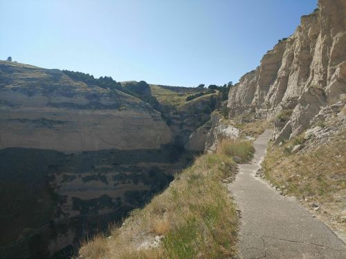 A narrow path winds along a rocky cliffside, with steep canyon walls and greenery under a clear blue sky.