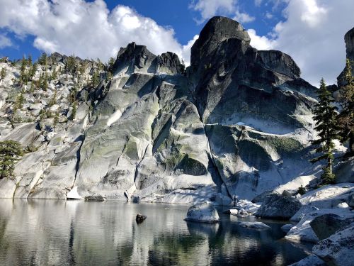 Majestic rocky cliffs reflect in a serene lake, surrounded by lush trees and a bright blue sky with fluffy clouds.