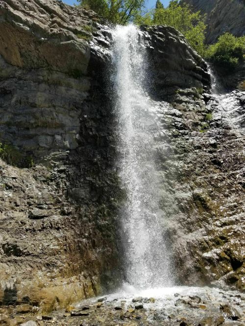A cascading waterfall flows down rocky cliffs, surrounded by lush greenery under a clear blue sky.