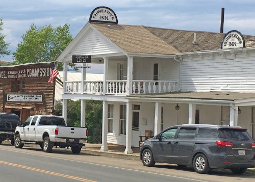 A white two-story inn with a porch, parked cars, and a nearby sign for a blacksmith shop in a small town.