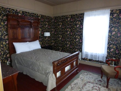 Cozy bedroom with a wooden bed, floral wallpaper, a window with sheer curtains, and a vintage armchair.