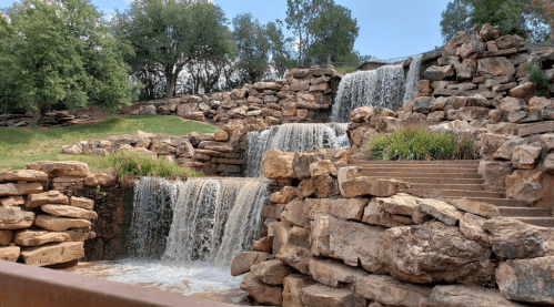 A serene waterfall cascading over rocky formations, surrounded by greenery and trees under a clear blue sky.