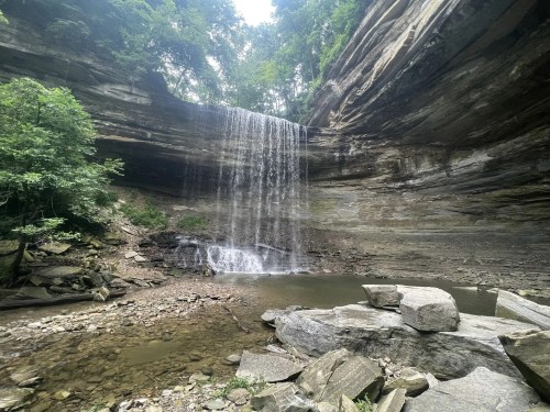 A serene waterfall cascades over rocky cliffs into a calm pool, surrounded by lush greenery and natural stone formations.
