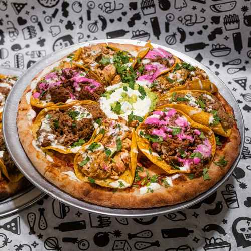 A pizza topped with various tacos, garnished with cilantro and a dollop of sour cream, on a patterned background.