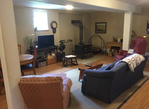 Cozy basement living area with a TV, seating, a wood stove, and a small dining table. Warm, inviting atmosphere.