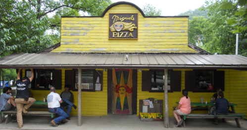 A yellow wooden building with a sign reading "Miguel's Pizza," featuring outdoor seating and patrons enjoying their meals.