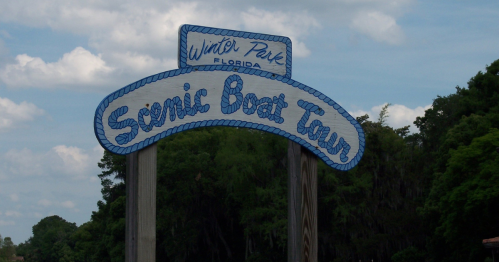 Sign for "Scenic Boat Tour" in Winter Park, Florida, with a blue rope design against a cloudy sky and green trees.