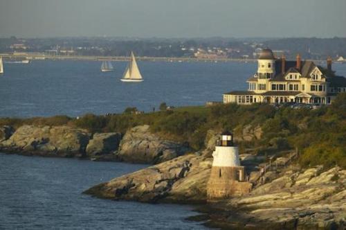 A coastal scene featuring a lighthouse, rocky shore, and sailboats on the water, with a large house in the background.
