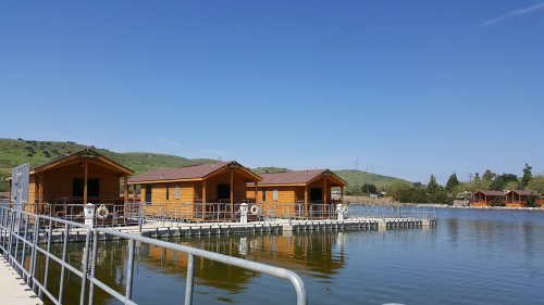 Three wooden cabins on a lake, connected by a walkway, surrounded by green hills under a clear blue sky.
