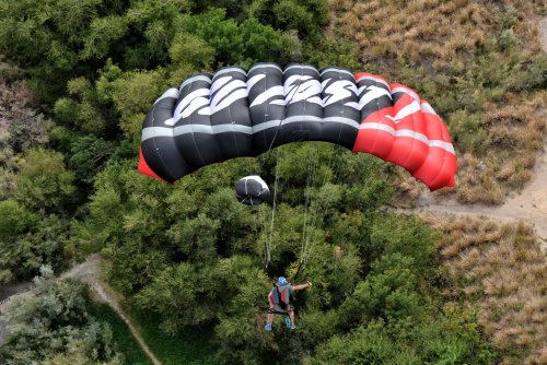 A person parachuting with a black and red parachute over a green landscape.