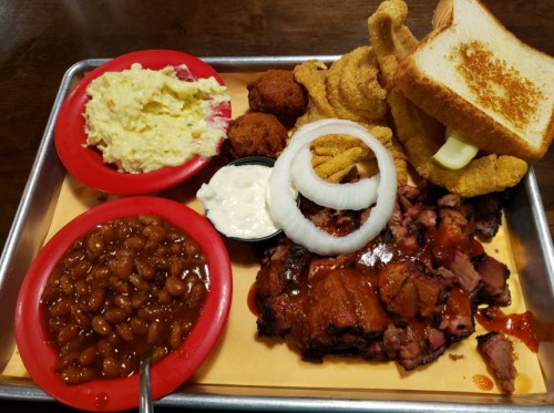 A platter of barbecue with brisket, baked beans, coleslaw, cornbread, onion rings, and toast on a tray.