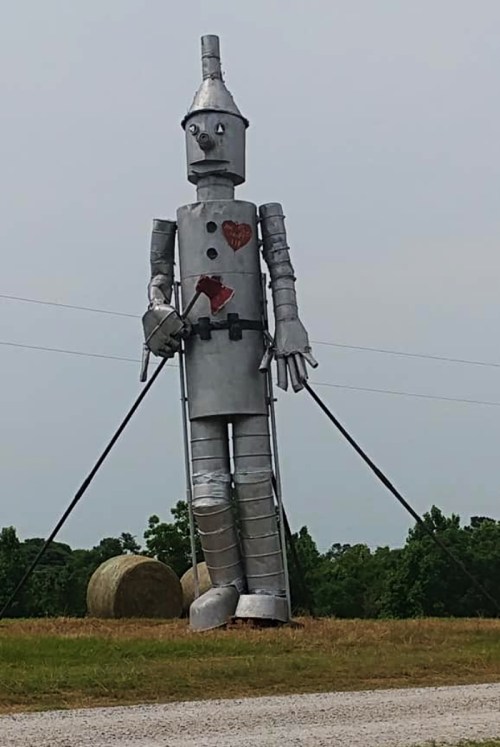 A large metal statue of a tin man holding an axe, standing in a grassy field with trees in the background.