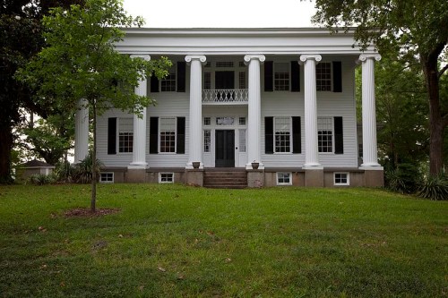 A large, two-story house with white columns, green lawn, and a symmetrical facade, surrounded by trees.
