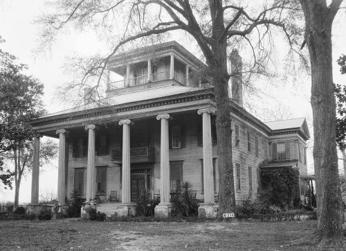 Historic two-story house with columns, a balcony, and large trees in the foreground, captured in black and white.
