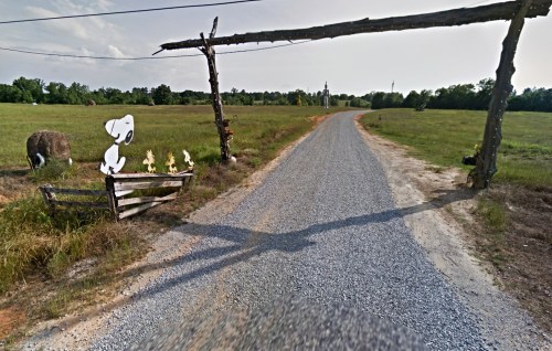 A gravel road leads through a rustic wooden archway, with cartoon characters and a hay bale on the side.