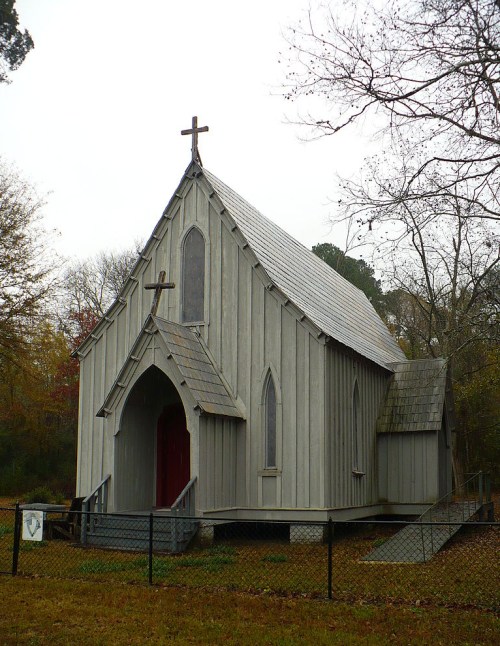 A gray wooden church with a steep roof, crosses on top, and surrounded by trees and a fence.