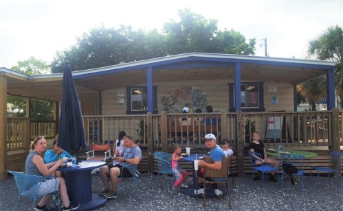 A group of people enjoying food and drinks outside a colorful café with a wooden deck and palm trees.