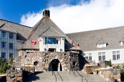 Historic stone building with flags, featuring a triangular roof and steps leading to the entrance, surrounded by greenery.