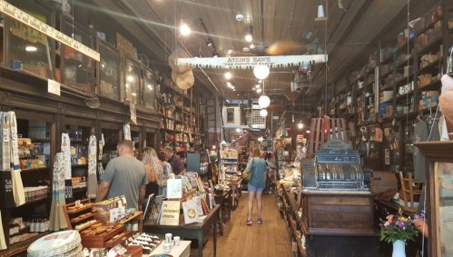 A vintage store interior filled with shelves of goods, people browsing, and an old cash register in the foreground.