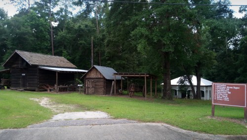 Historic wooden buildings surrounded by trees, with a sign listing various attractions and amenities nearby.