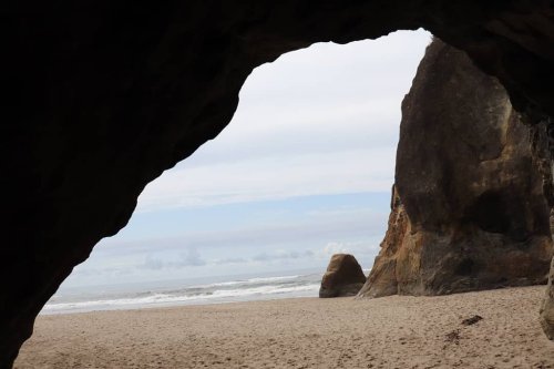 View of a sandy beach framed by a rocky arch, with waves gently lapping at the shore under a cloudy sky.