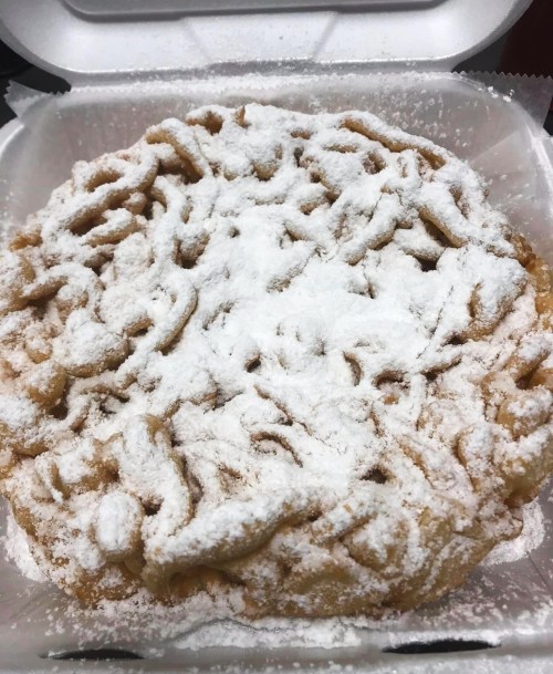 A freshly made funnel cake dusted with powdered sugar, served in a takeout container.