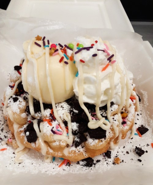 A delicious funnel cake topped with ice cream, whipped cream, sprinkles, and crushed cookies, served in a takeout container.