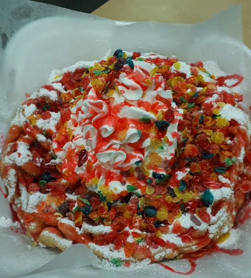 A colorful funnel cake topped with whipped cream, fruit syrup, and a mix of fruity cereal pieces.
