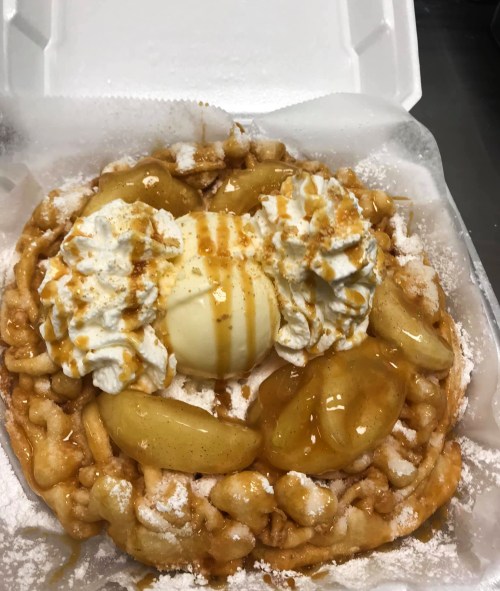 A delicious funnel cake topped with whipped cream, ice cream, caramel, and warm apple slices, served in a takeout container.